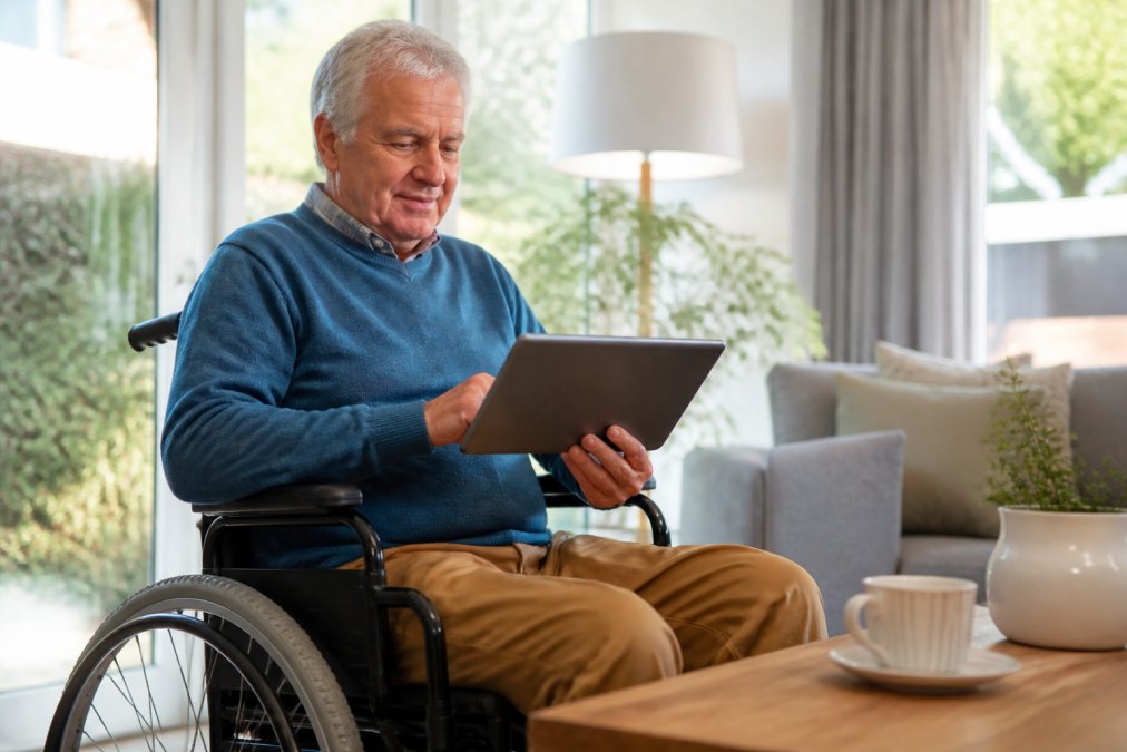 Oudere persoon in rolstoel die op een tablet zit en online een rit reserveert, met een vriendelijke, gerichte blik op het scherm in een zonnig interieur