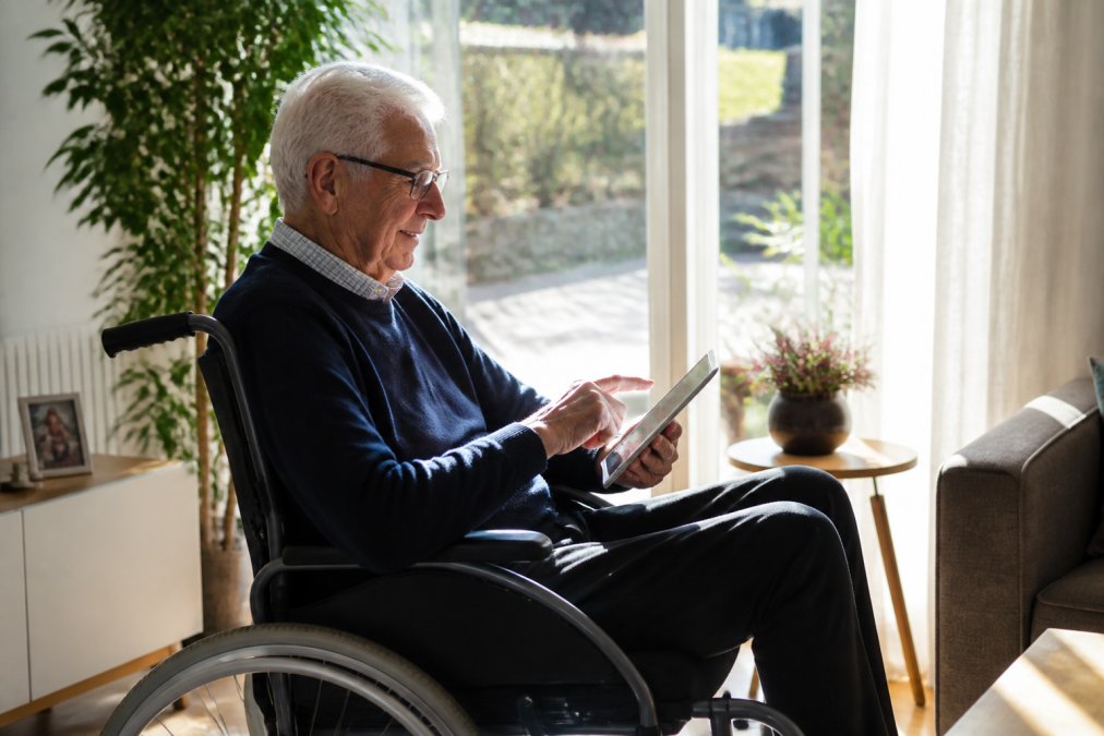 Oudere persoon in een rolstoel reserveert online een rolstoeltaxi 24 uur via een tablet in een zonnig interieur.