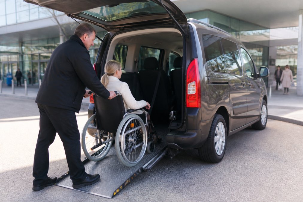Een vriendelijke, medisch gecertificeerde chauffeur helpt voorzichtig een rolstoelgebruiker in een speciaal aangepaste rolstoeltaxi met open deuren en duidelijk zichtbare aangepaste ingang, op een zonnige ochtend in Utrecht