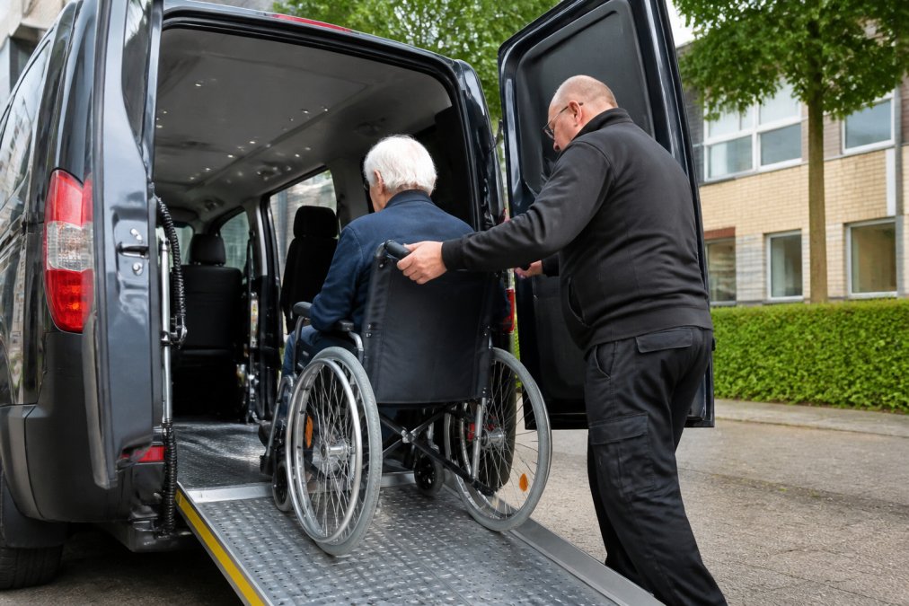 Getrainde chauffeur helpt een rolstoelgebruiker veilig in een voertuig met oprijplaat, toont professionele en zorgzame bediening in helder daglicht