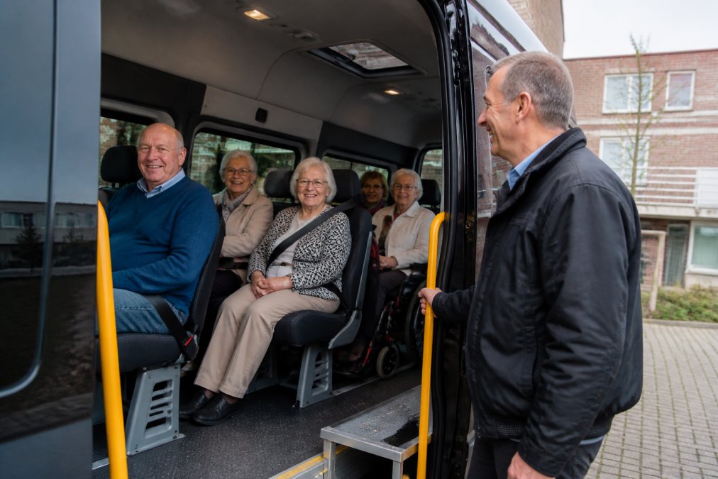 Groep senioren en cliënten met verschillende mobiliteitsbehoeften die samen in een ruime, goed uitgeruste vervoersbus zitten met zachte zitplaatsen en veiligheidsfuncties; dezelfde chauffeur staat bij de deur
