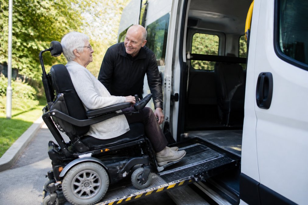 Oudere vrouw in elektrische rolstoel wordt geholpen door vriendelijke mannelijke chauffeur bij het instappen in rolstoeltaxi, met zichtbare oprijplaat en open deur, daglicht van buiten