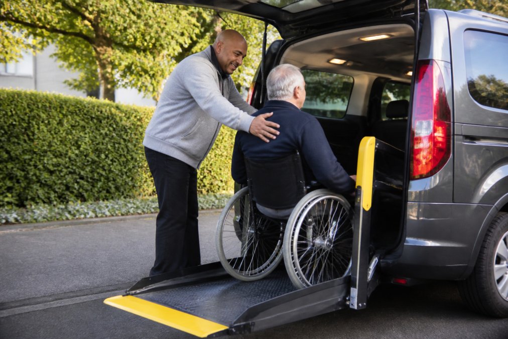 Een vriendelijke, ervaren chauffeur helpt een rolstoelgebruiker stap voor stap bij het instappen in een moderne, schone rolstoeltaxi met een geopende lift, terwijl de chauffeur een geruststellend gebaar maakt
