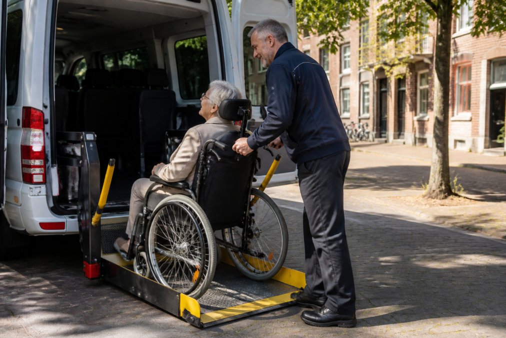 Een oudere mevrouw in een rolstoel wordt door een vriendelijke chauffeur in uniform veilig geholpen bij het instappen in een wit busje met rolstoellift, op een rustige straat in Utrecht bij daglicht.
