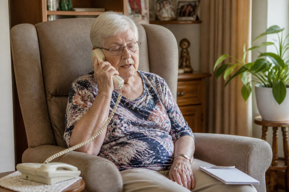 Een oudere persoon zit thuis in een comfortabele stoel en belt telefonisch om een rolstoeltaxi te reserveren, met een notitieblokje naast zich voor de reisinformatie