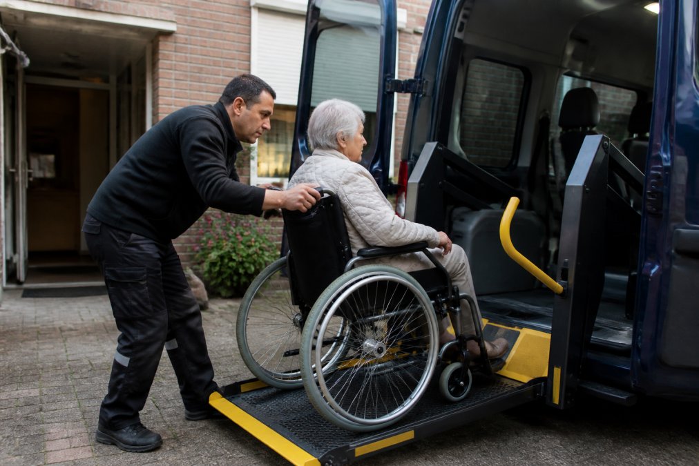 Een medisch gecertificeerde chauffeur helpt een senior in een rolstoel voorzichtig in een rolstoeltaxi in te stappen, met beide handen ondersteunend, in daglicht buiten een woning