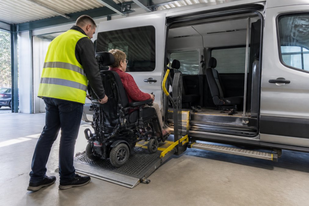 Aangepaste rolstoeltaxi met volledig open zijdeur, hydraulische lift in werking, chauffeur met reflectiehesje helpt cliënt met elektrische rolstoel voorzichtig in voertuig, comfortabel interieur met bevestigingssystemen zichtbaar, daglicht in moderne garage