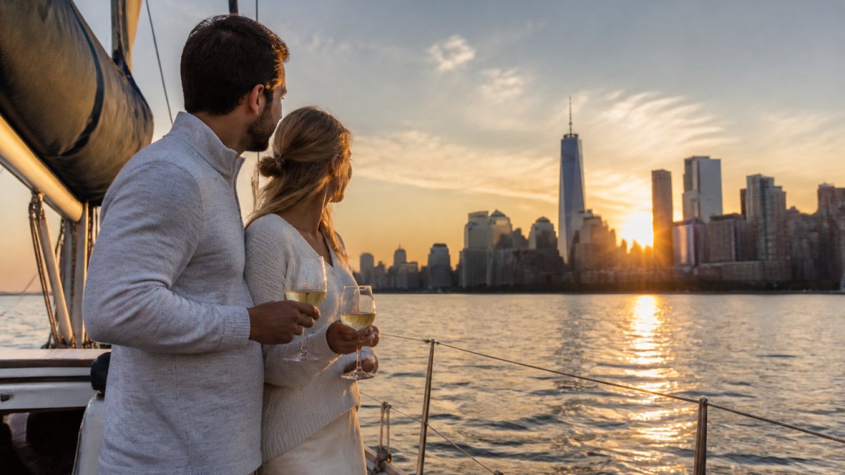A couple standing on the deck of a private sailboat at sunset, holding glasses of wine, with the Manhattan skyline glowing in the background and the water reflecting golden light