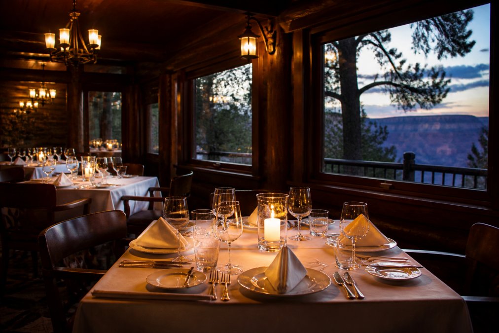 An elegantly set dinner table inside El Tovar's historic lodge dining room, with warm dark wood paneling, candlelight reflecting off wine glasses, white linen tablecloths, and a window hinting at the canyon's rim just outside in the fading evening light