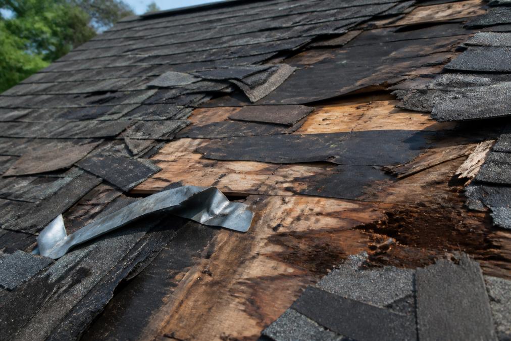 Close-up detail of severe roof damage showing multiple missing shingles, exposed dark underlayment, bent metal flashing, and water-stained wood substrate on residential roof