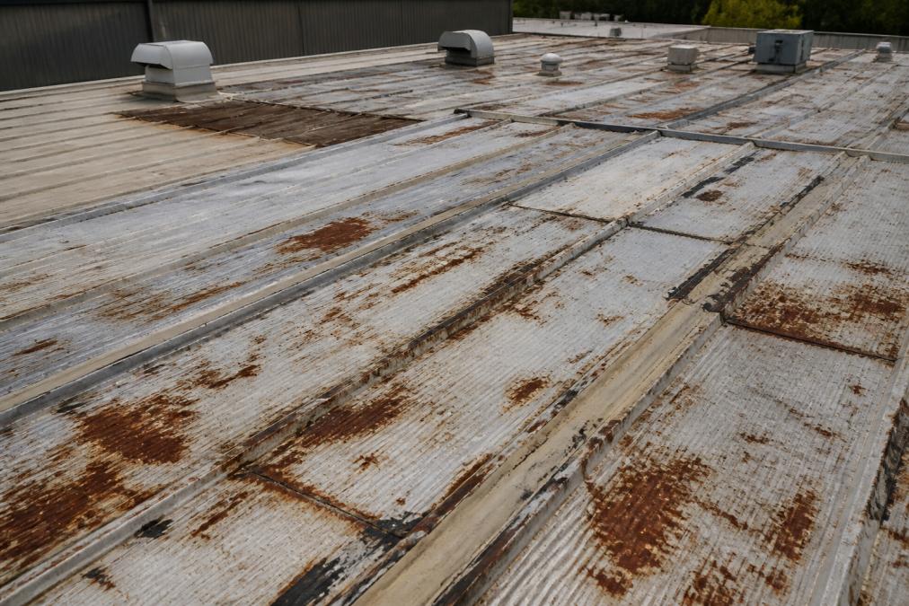 Overhead view of a commercial industrial roof with visible rust patches, deteriorating metal panels, and worn seams showing the condition that triggers refurbishment decisions