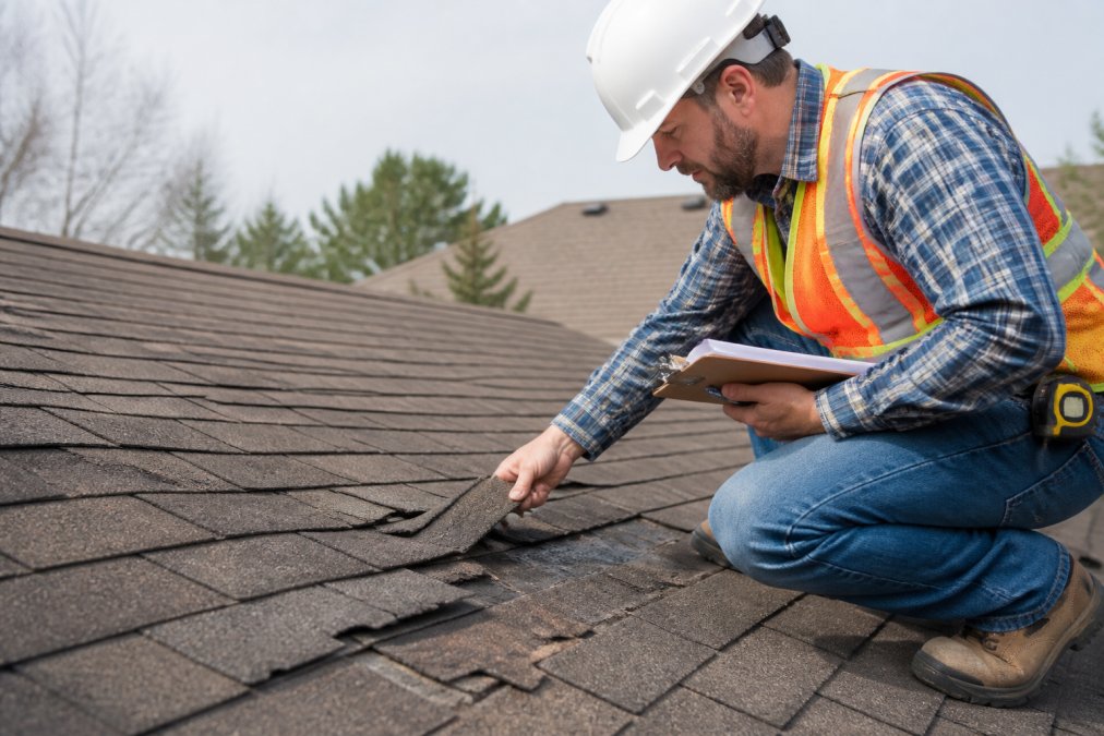 Professional roofing contractor in safety gear and hard hat inspecting a residential roof with clipboard and measuring tape, examining shingle condition and structural integrity