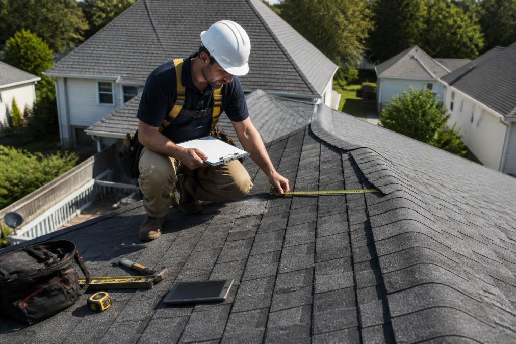 Professional roofing contractor in safety harness and hard hat performing detailed aerial inspection of residential roof with measurement tools and clipboard