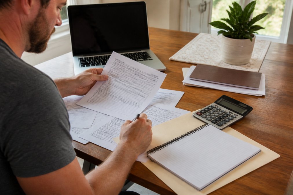 Homeowner at a wooden desk reviewing detailed loan paperwork with a laptop and calculator visible, preparing to submit roof replacement financing application with organized documents