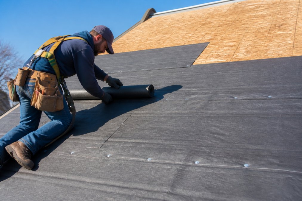 Professional roofer installing underlayment on roof deck, showing proper overlap technique and fastening pattern with visible nail placement