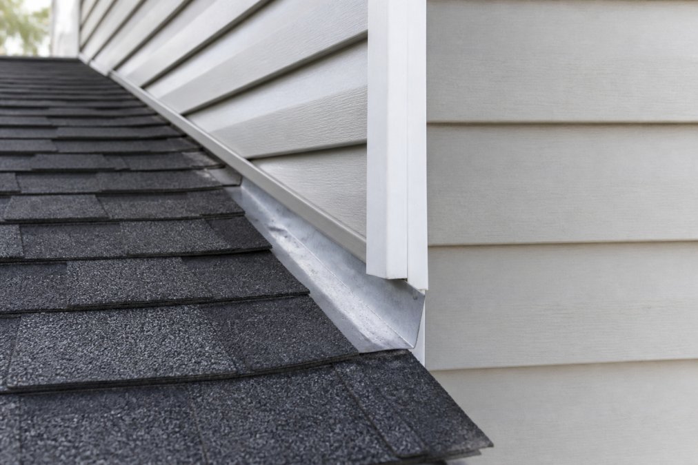 Close-up of residential home's roof-to-wall junction showing metal flashing, j-moulding trim, and where asphalt shingles meet the top of vinyl siding under clear daylight