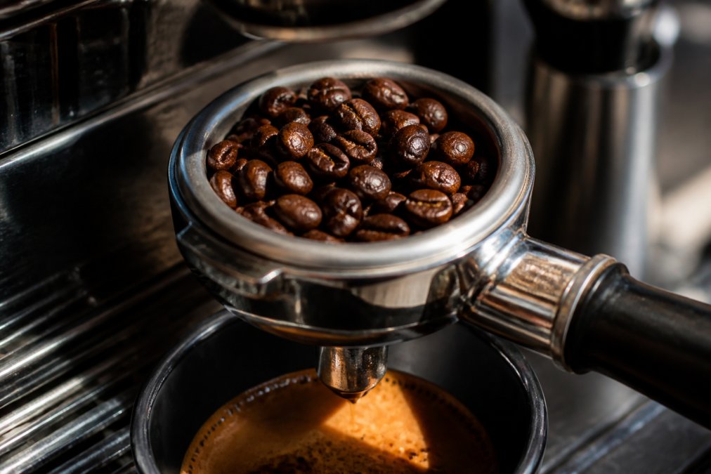 Close-up of dark espresso beans in a metal portafilter with golden crema pooling beneath, morning light highlighting the glossy surface of freshly roasted beans