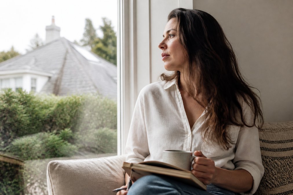 A woman sitting quietly by a window with a journal and a cup of tea, soft natural light, a calm reflective expression, conveying the moment of noticing a familiar emotional pattern without self-judgement