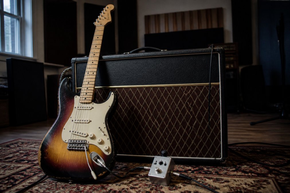 A vintage sunburst Stratocaster leaning against a classic Vox AC30 amplifier in a dimly lit music studio, with a treble booster pedal on the floor in front