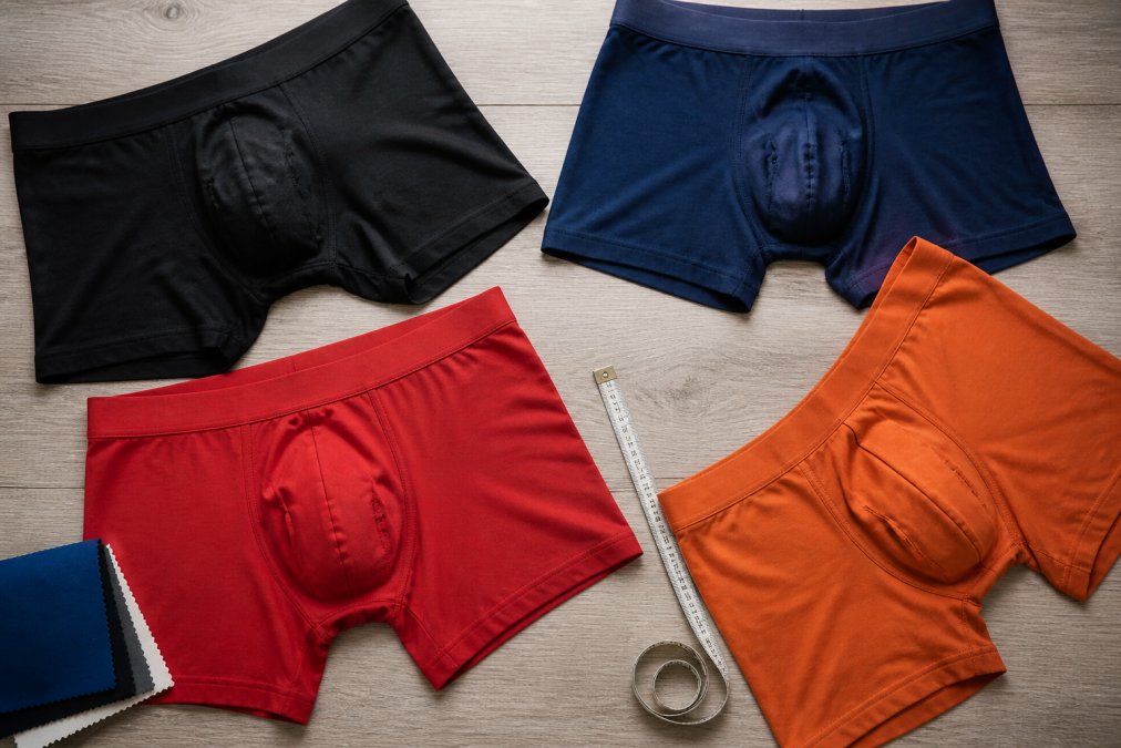 Flat lay of men's boxer briefs in black, navy, red, and orange arranged on a light wood surface with a measuring tape and fabric swatch nearby, showing pouch construction detail