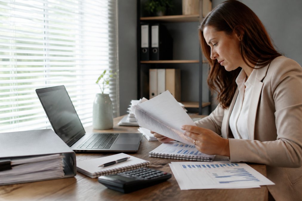 A recruitment manager reviewing candidate files beside a laptop showing a hiring cost spreadsheet at a desk in a bright office
