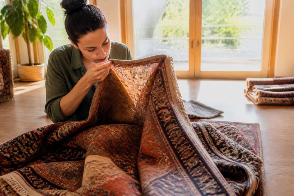 Person carefully examining a vintage rug's weave and pile construction in a bright home interior, holding fabric up to natural light to inspect fiber quality and dye characteristics