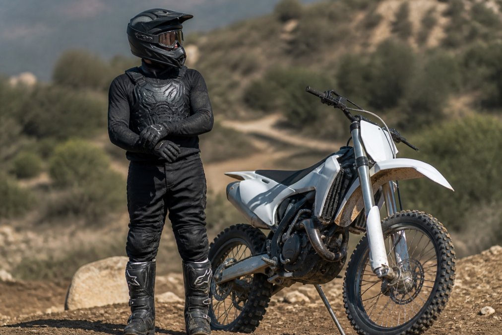 Rider in full protective gear standing next to dirt bike displaying helmet, goggles, chest protector, gloves, and boots in outdoor trail setting with natural lighting
