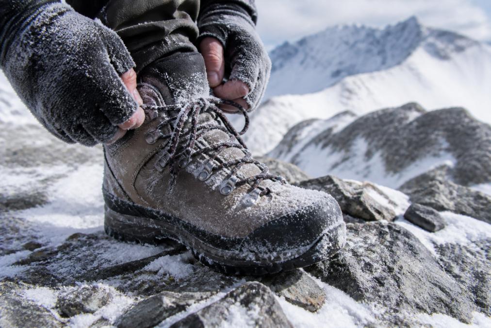 Close-up of rugged waterproof hiking boots secured tightly on rocky, snow-covered mountain terrain with frost-covered laces and hands adjusting the boot collar
