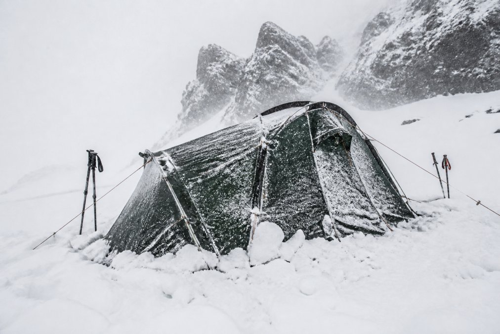 A four-season tent set up in heavy, accumulating snow with frost-covered fabric, guy lines taut under wind load, showing weatherproof construction and reinforced poles in sub-zero conditions