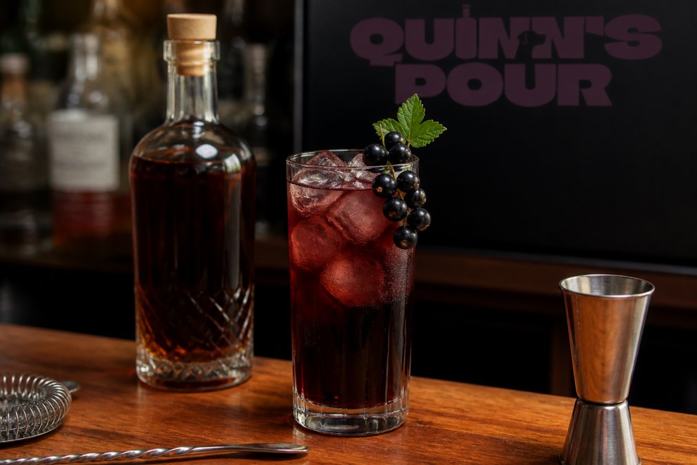 A highball glass filled with dark rum and blackcurrant cordial over ice, garnished with a blackcurrant sprig, placed on a wooden home bar counter with a bottle of premium spiced rum beside it