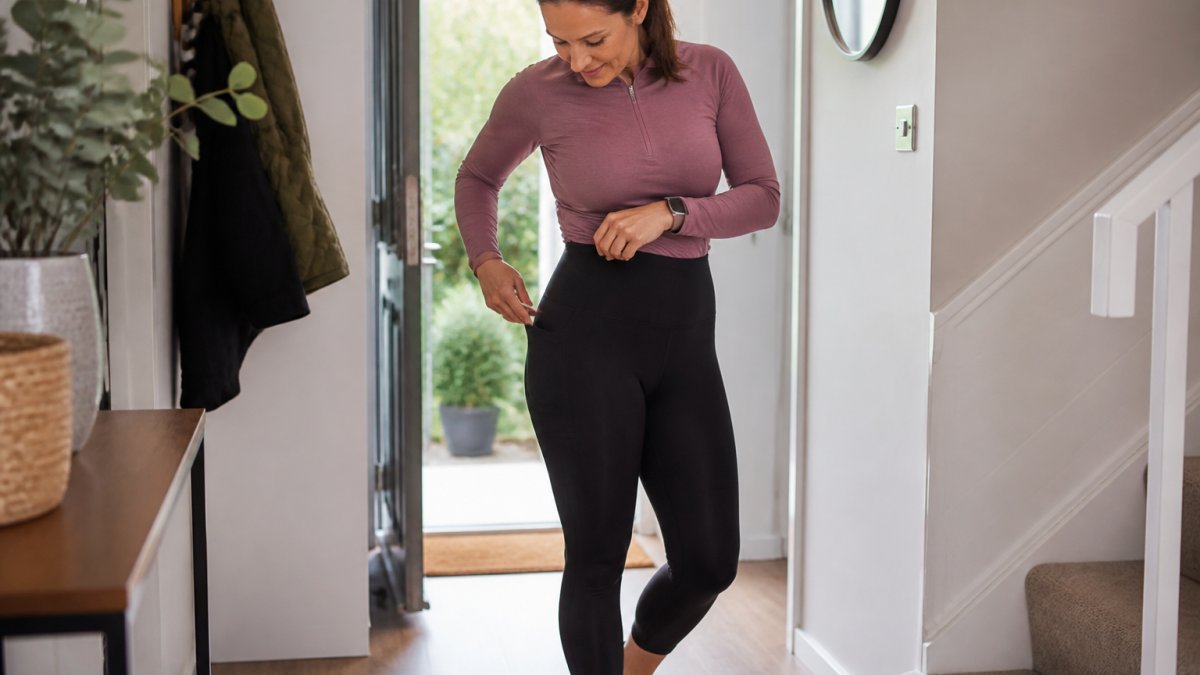 A woman in her forties wearing high-waisted black running leggings, checking the waistband and pocket detail in a bright home hallway before heading out for a run
