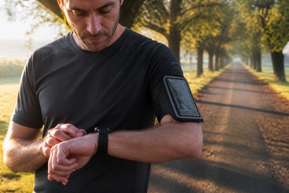 A runner checking a GPS watch on their wrist while a smartphone sits in an armband, shot outdoors on a tree-lined path in soft morning light