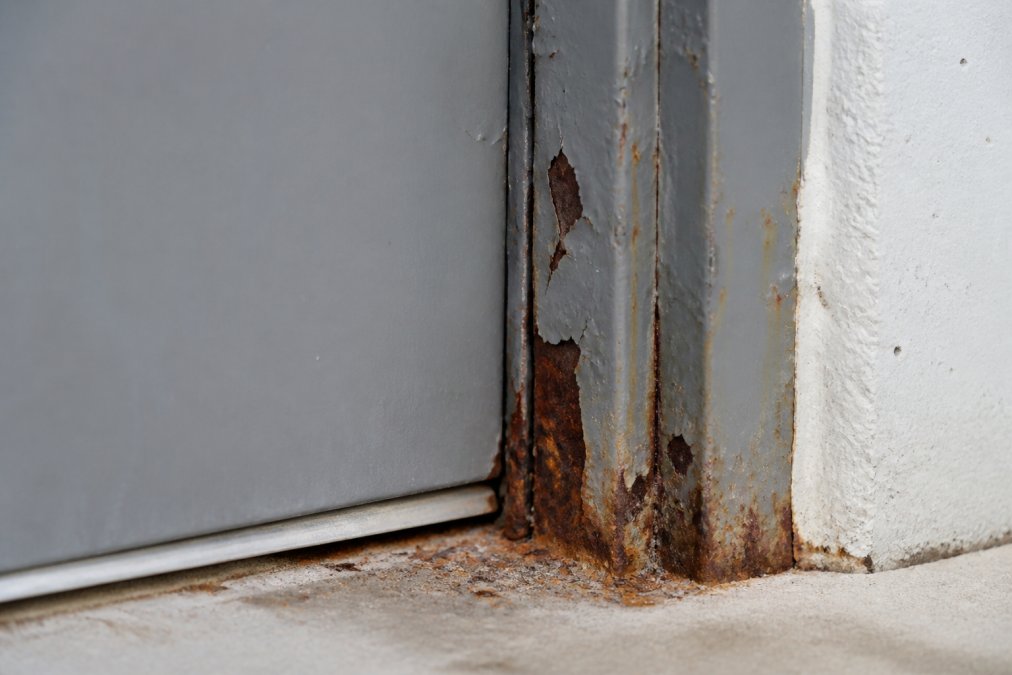 Close-up of commercial steel door frame showing visible rust stains, flaking paint, and orange-brown corrosion at the base and jamb areas in natural daylight
