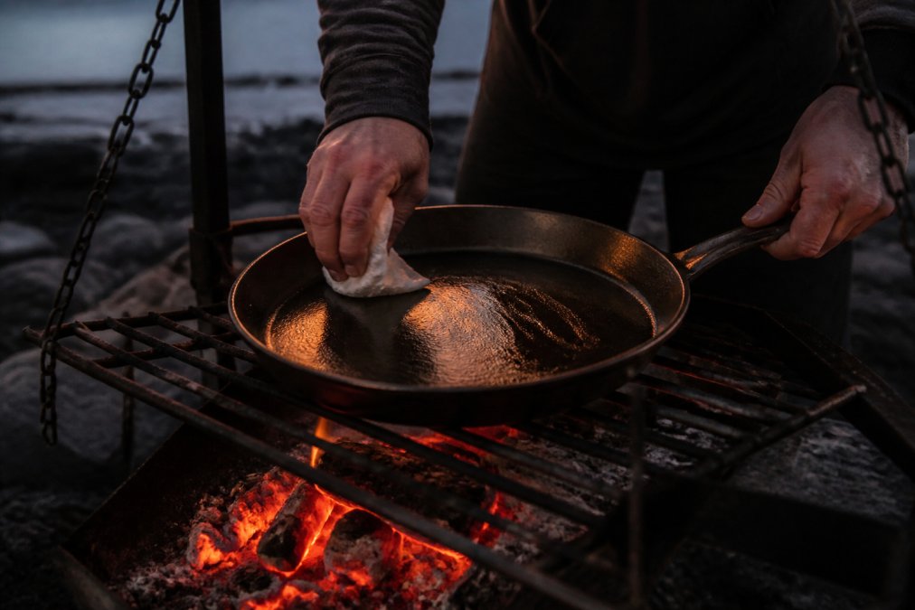 Close-up of hands applying a thin layer of oil to a cast iron skillet over glowing campfire coals at dusk, showing the golden-brown seasoning patina