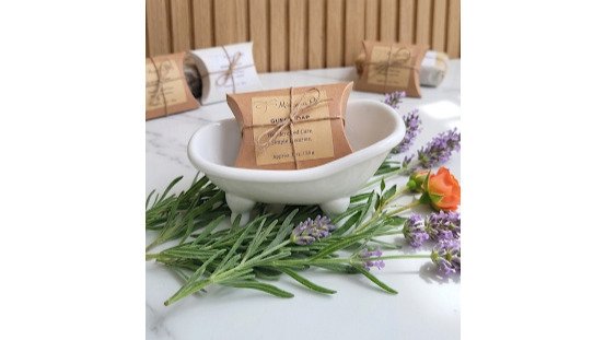 Overhead flat-lay arrangement of handcrafted artisanal soap bars in rustic kraft pillow boxes with natural twine bows, displayed on neutral linen with dried lavender sprigs and fresh citrus slices, natural window lighting