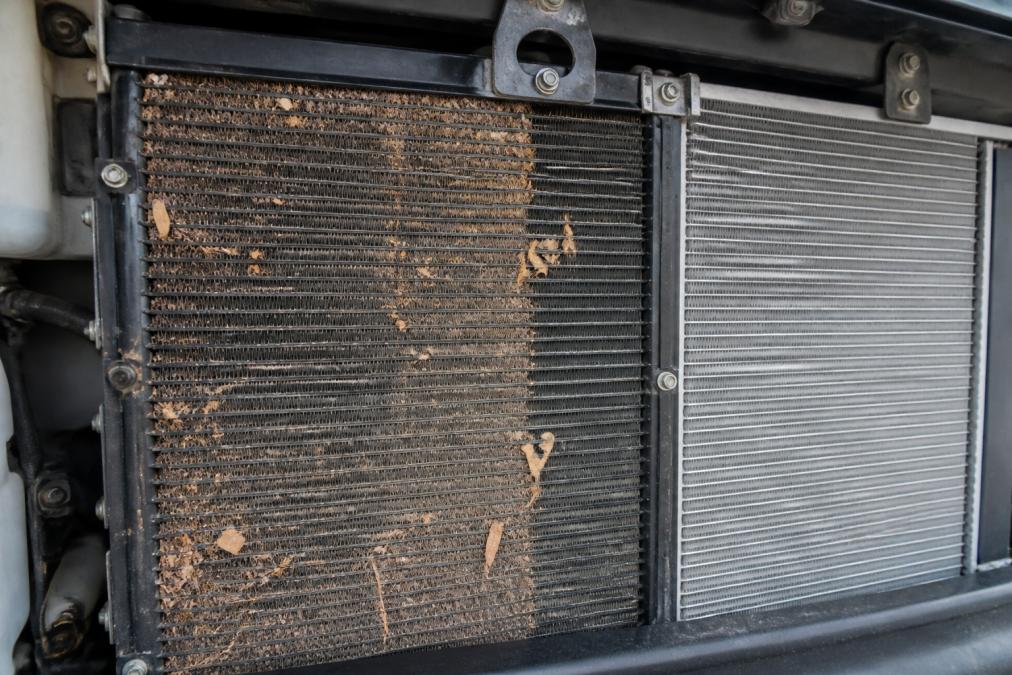 Close-up of a diesel RV radiator with visible dirt, leaves, and debris clogging the aluminum fins, showing the contrast between clean and blocked cooling surfaces under natural daylight