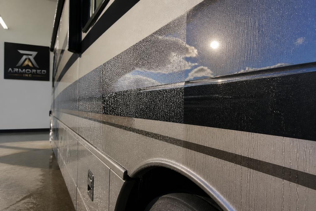 Close-up of an RV exterior surface with water beading on a freshly ceramic-coated panel, sunlight reflecting off the glossy finish, showing contrast between coated and uncoated areas