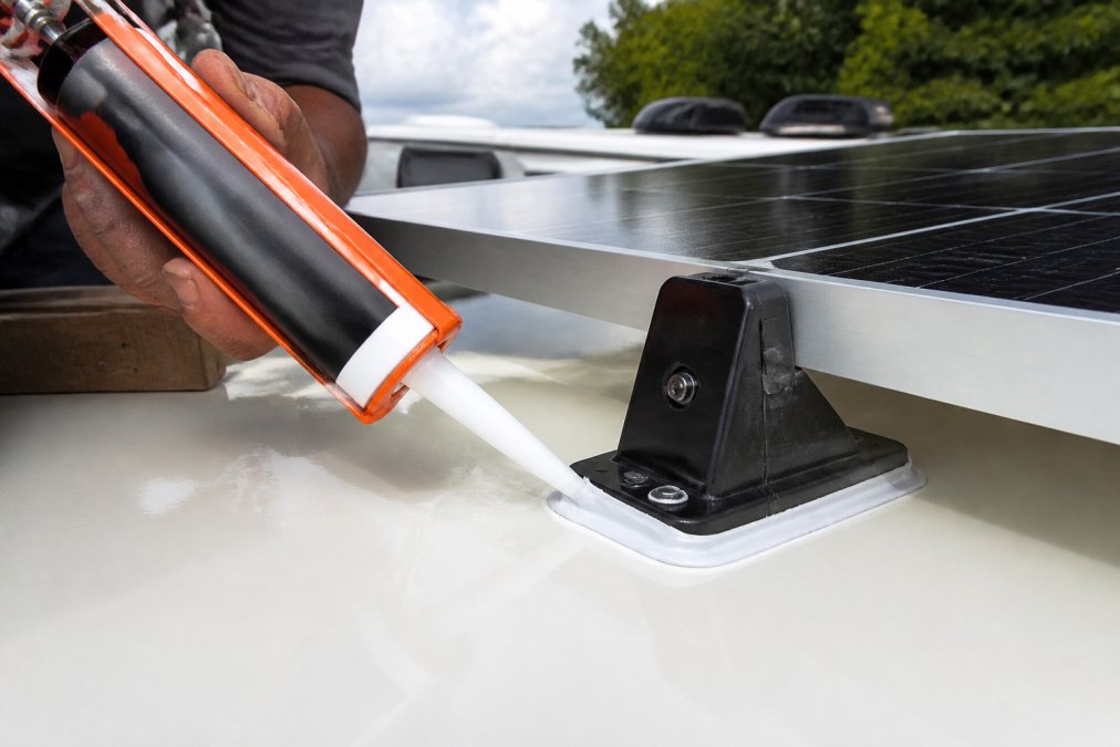 Technician applying marine-grade sealant to a solar panel mounting foot on an RV roof using a caulk gun.