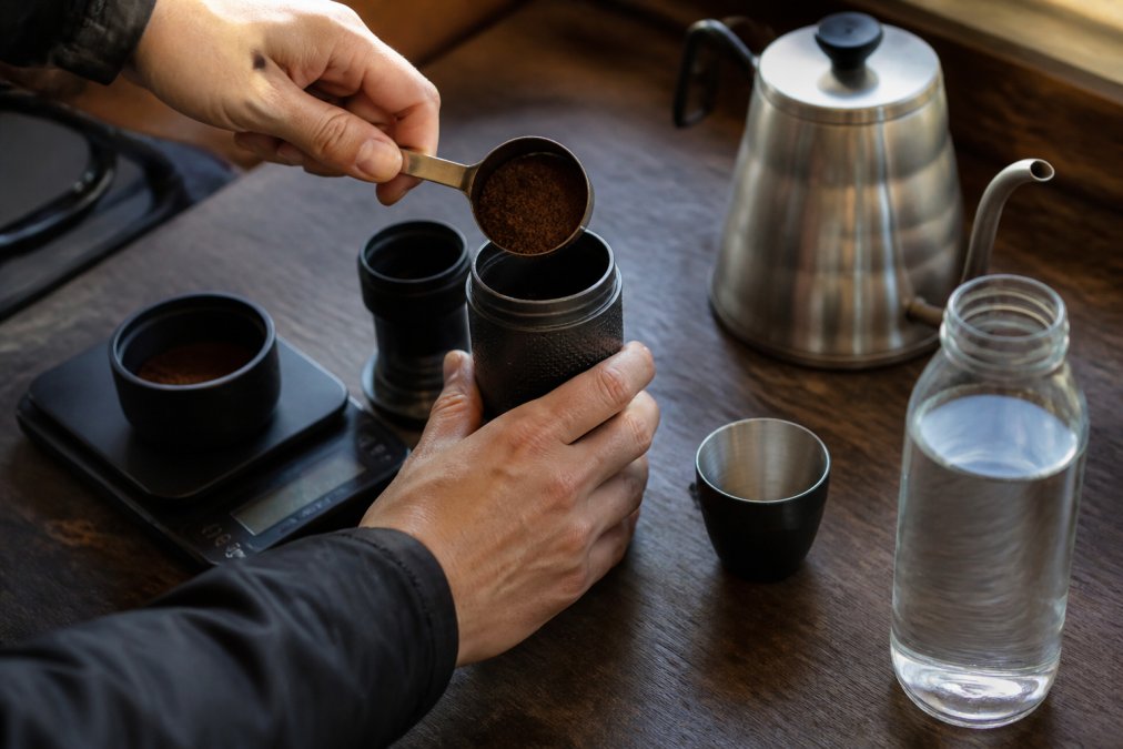 Hænder der måler og doserer fintmalede kaffebønner i en håndholdt espressopresser, med digitale vægt og målekop på træbord ved siden af varmt vand