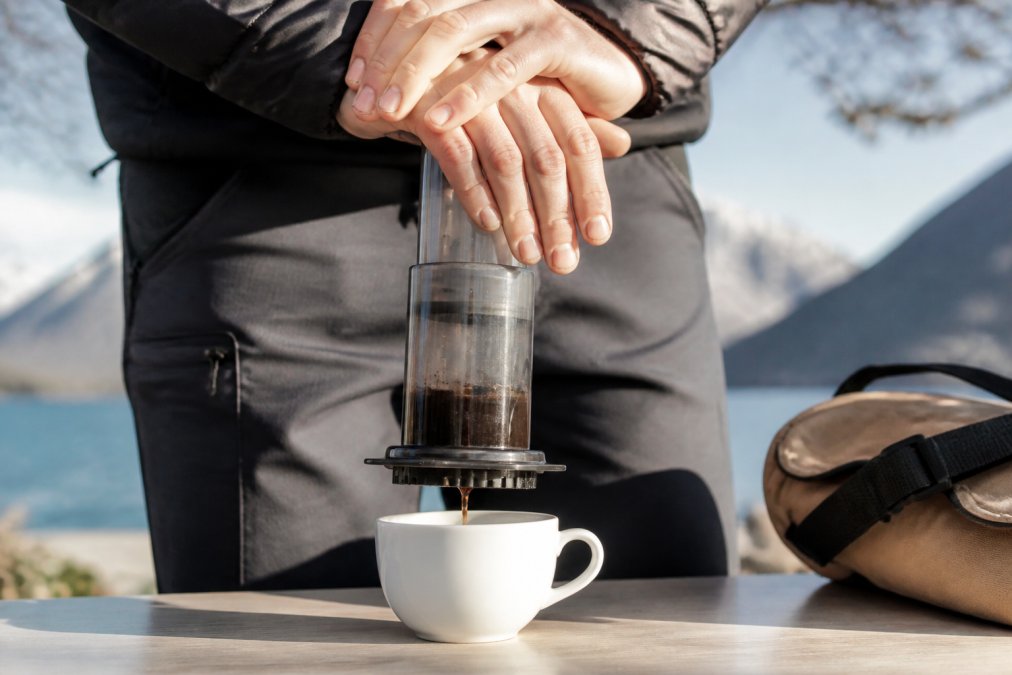 Hænder der demonstrerer presning af Aeropress over en hvid keramisk kop, med fokus på stemplets bevægelse og det mørke kaffegrums der langsomt kommer ud