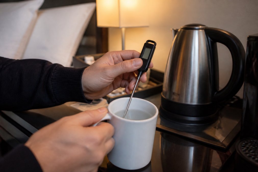 Nærbillede af hænder, der holder et lille digitalt termometer i en kop varmt vand på et hotelskrivebord med en elkedel i baggrunden