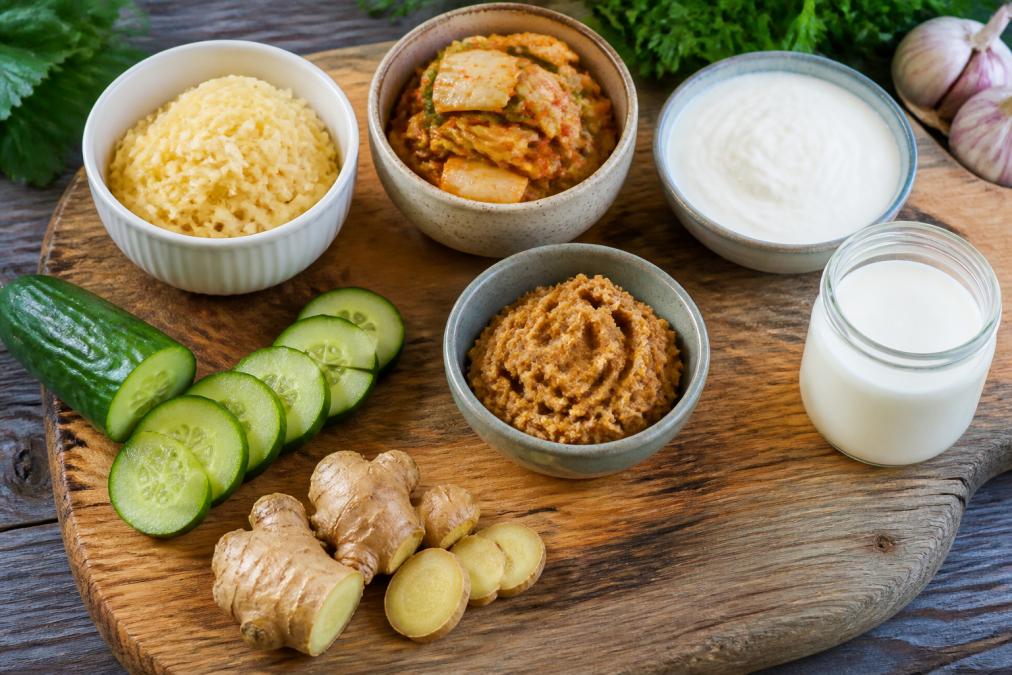 Overhead shot of wooden cutting board with fermented foods including bowls of sauerkraut, kimchi, yogurt, kefir, and miso paste, with fresh vegetables like cucumber and ginger nearby in natural daylight