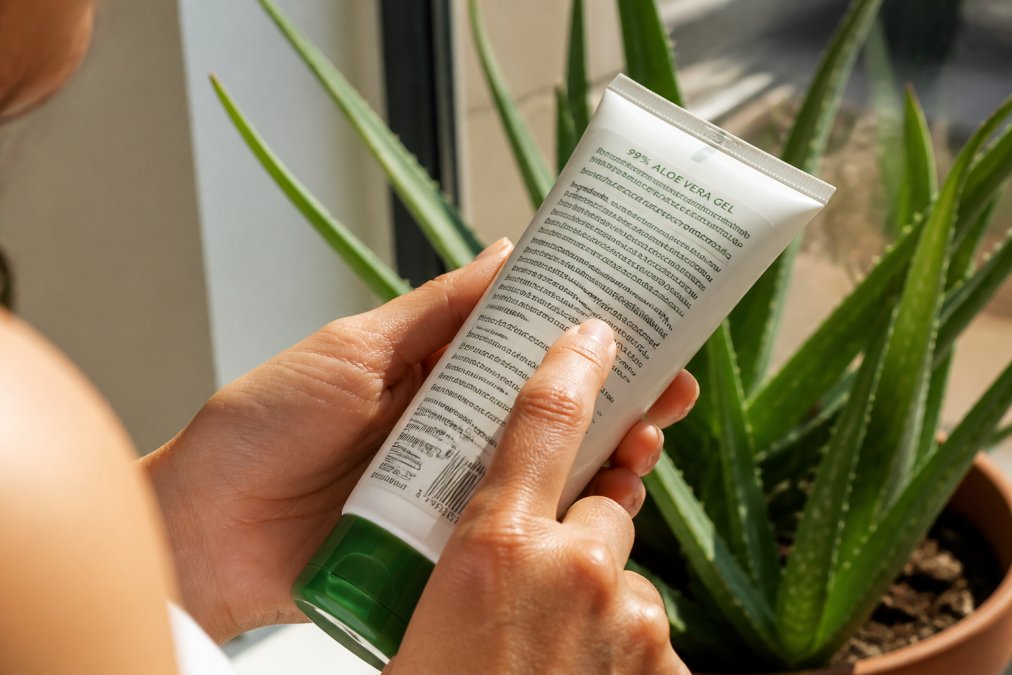 Hænder, der holder en aloe vera-produktflaske og læser ingredienslisten tæt under naturligt sollys, med etiketteksten klart synlig