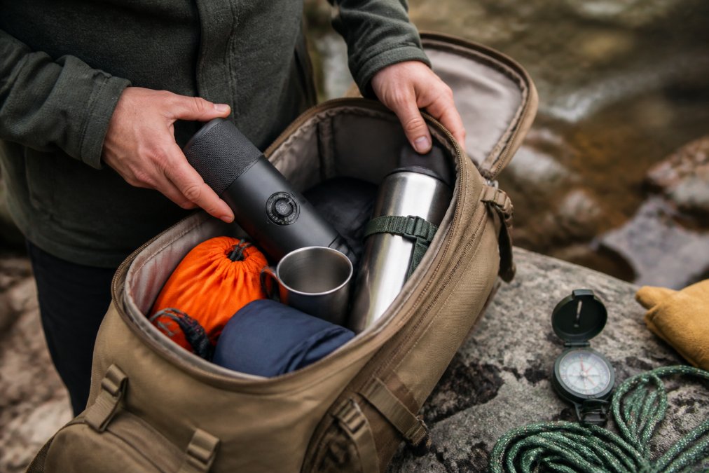 Close-up of a portable espresso machine being packed into a rugged backpack alongside camping gear, with a compass and a thermos visible in the frame