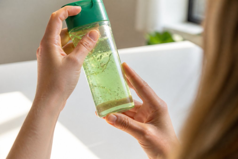 Tæt billede af hænder, der holder en flaske aloe vera gel op mod lyset for at undersøge farven og konsistensen, mens etiketten læses omhyggeligt på en hvid baggrund