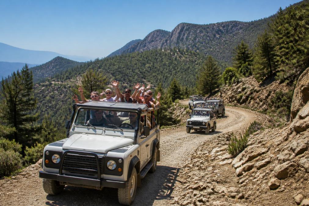 Jeep araçlarında güvenlik ekipmanları ile donanmış turist grubu, engebeli dağ arazisinde hareket halinde, yeşil orman arka planında ve açık mavi gökyüzü altında