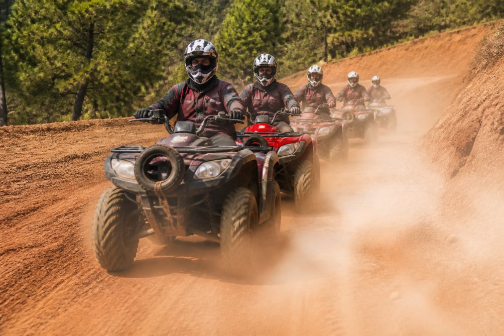 Quad araçlarında güvenlik ekipmanları (kask, eldiven, koruyucu ceket) ile donanmış turist grubu, kırmızı topraklı arazide hareket halinde, güneşli hava ve ön planda toz bulutu