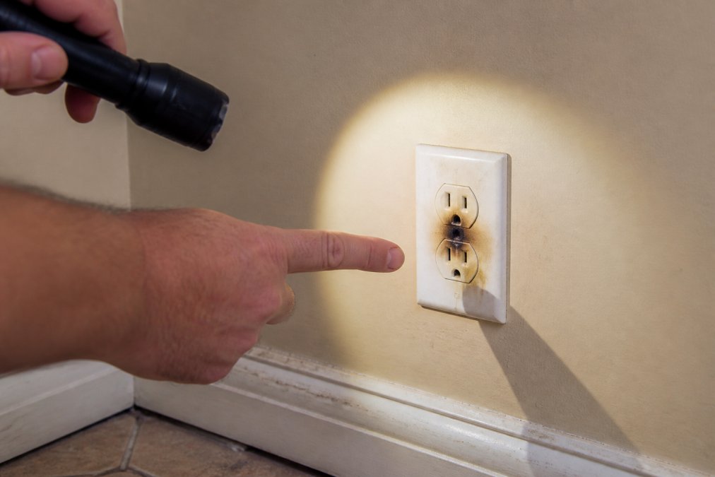 Close-up of a homeowner's hand pointing to a discolored, scorched electrical outlet on a beige wall, with a flashlight beam highlighting the burn marks