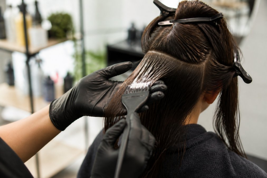 Close-up of hands applying keratin treatment to sectioned damp hair with clips holding sections in place, professional setting with natural lighting