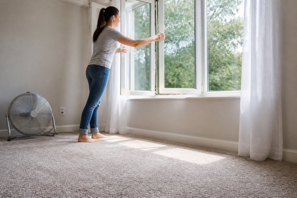 A parent opening windows in a home to allow air circulation, with a fan visible in the background and carpet visibly drying in natural light